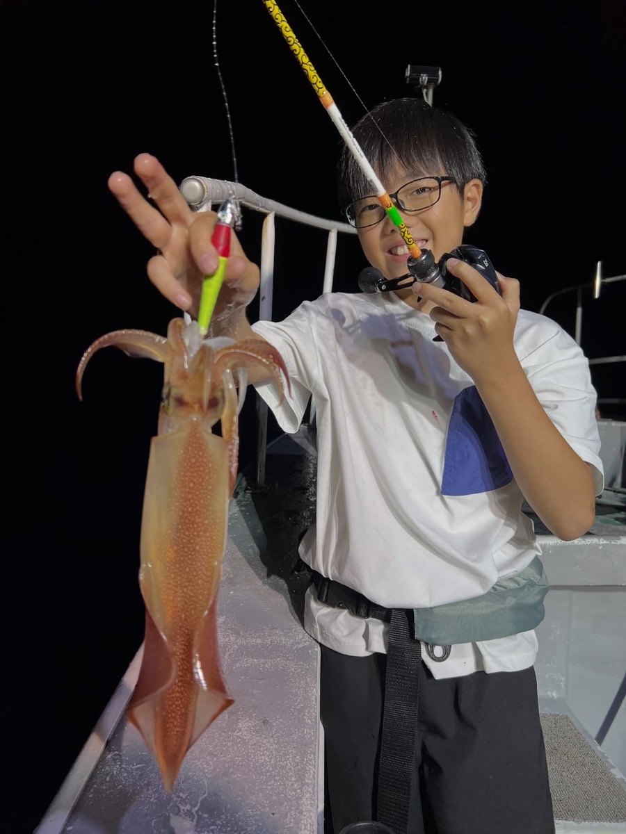 夜の船上で、眼鏡をかけた少年がマイカとイカメタルタックルを掲げている