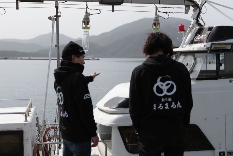 船の上で話す船長2人。奥に集魚灯と山が見える