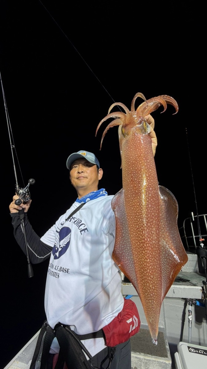 夜の船上で、釣り上げた大きなマイカを右手に掲げ、左手に竿を持つ男性
