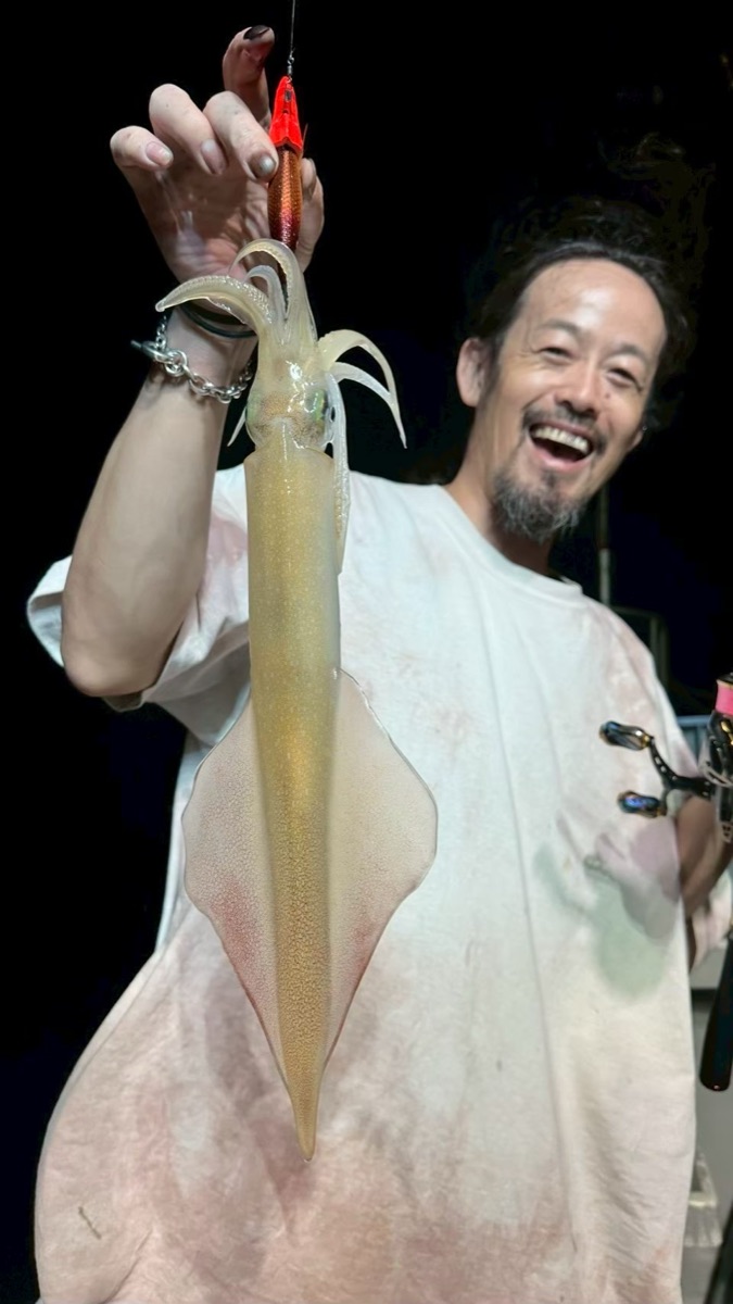 夜の船上で、白いシャツの男性が釣り上げたマイカを笑顔で掲げている