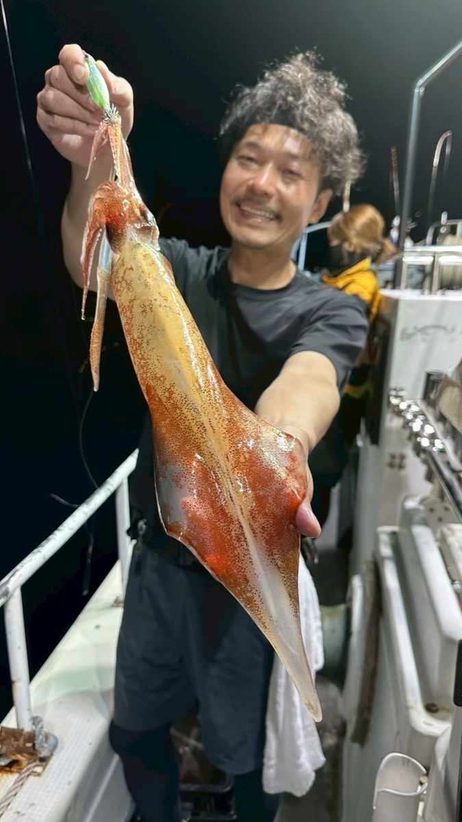 夜の船上で、男性が釣り上げたマイカをカメラへ差し出して笑っている