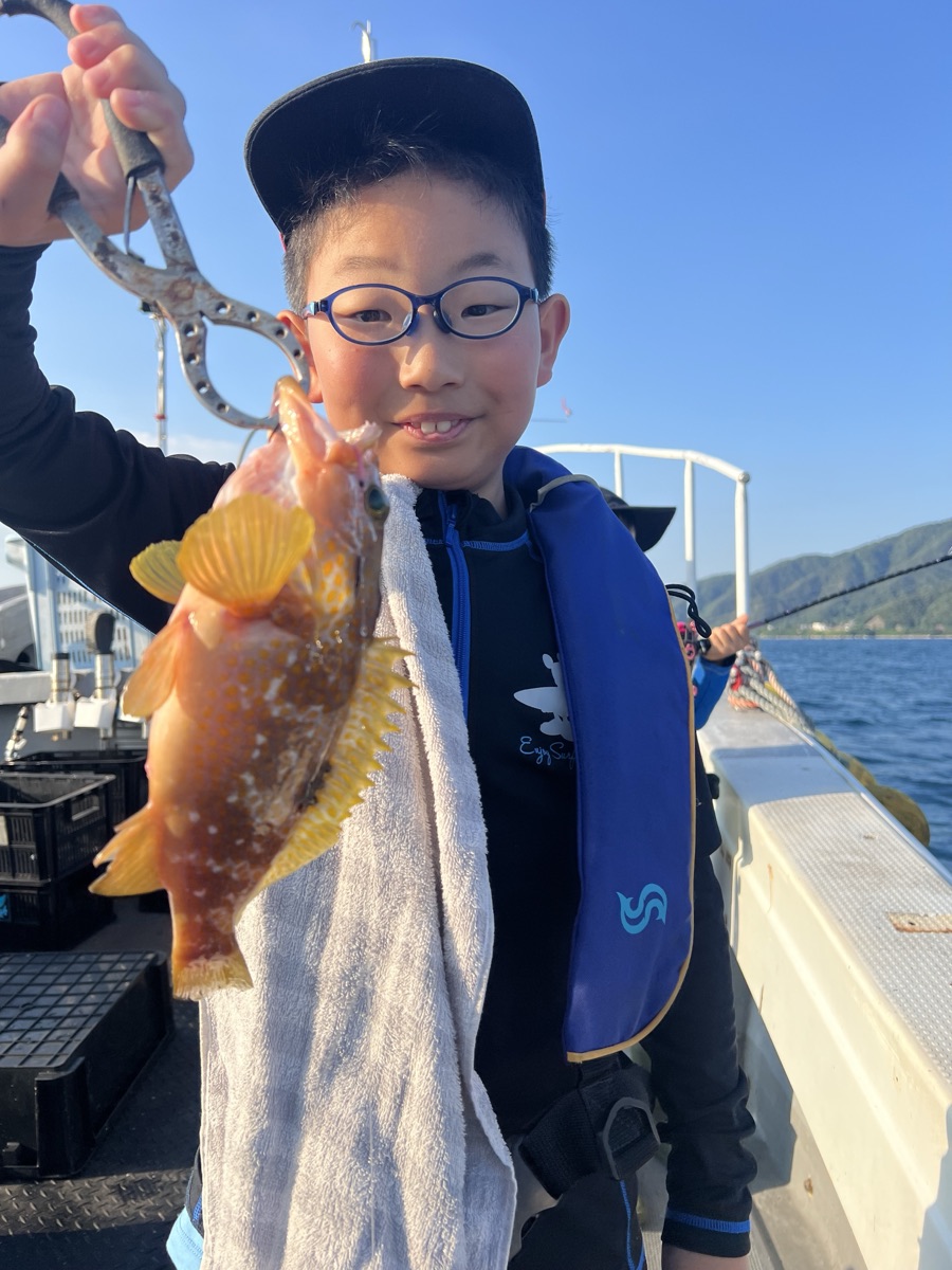 青空の船上で、眼鏡をかけた男の子が釣り上げた橙色の魚を掲げている