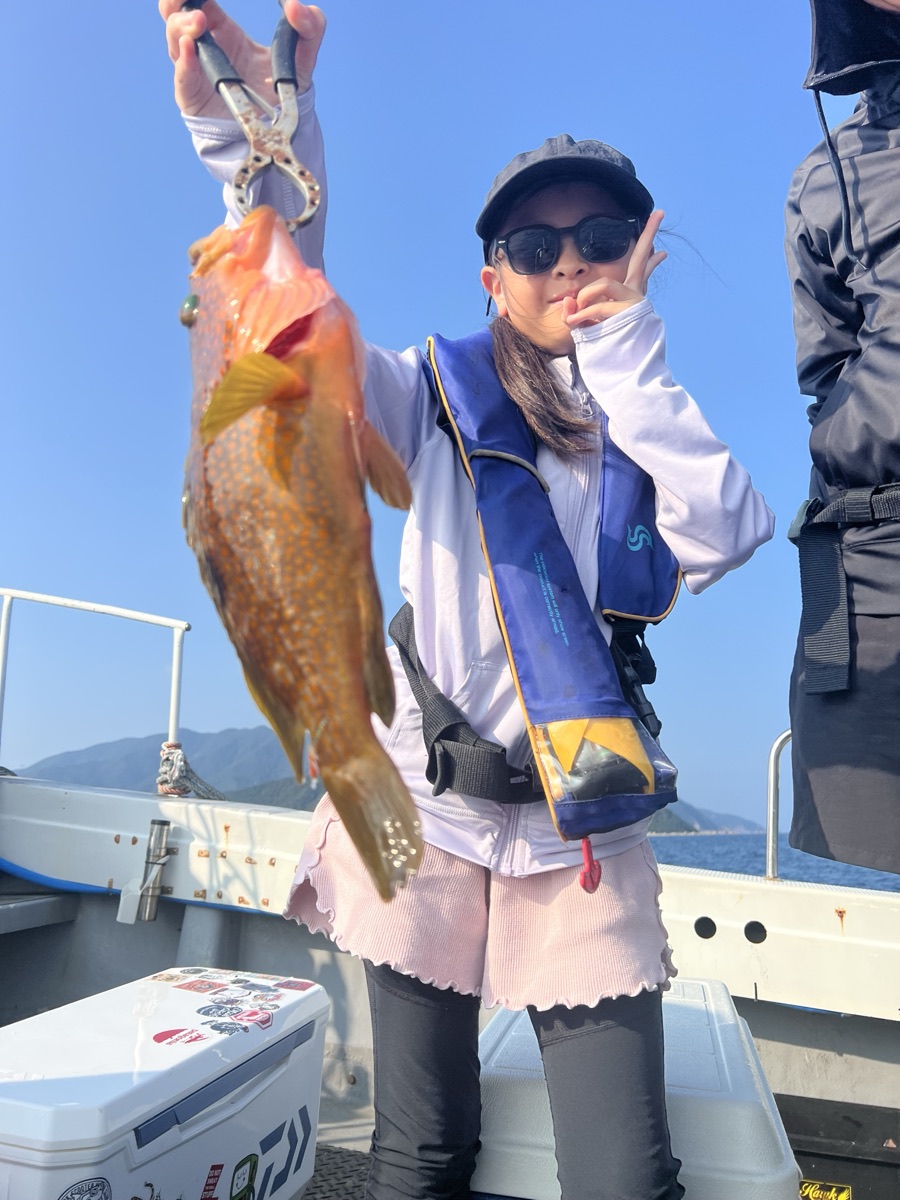 青空の船上で、サングラスをかけた女の子が釣り上げた橙色の魚を掲げている
