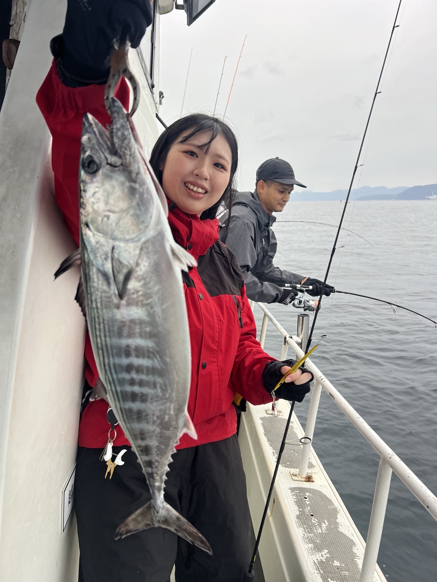 曇り空の船上で、赤い雨具の女性が縞模様の魚とジグを掲げて笑っている