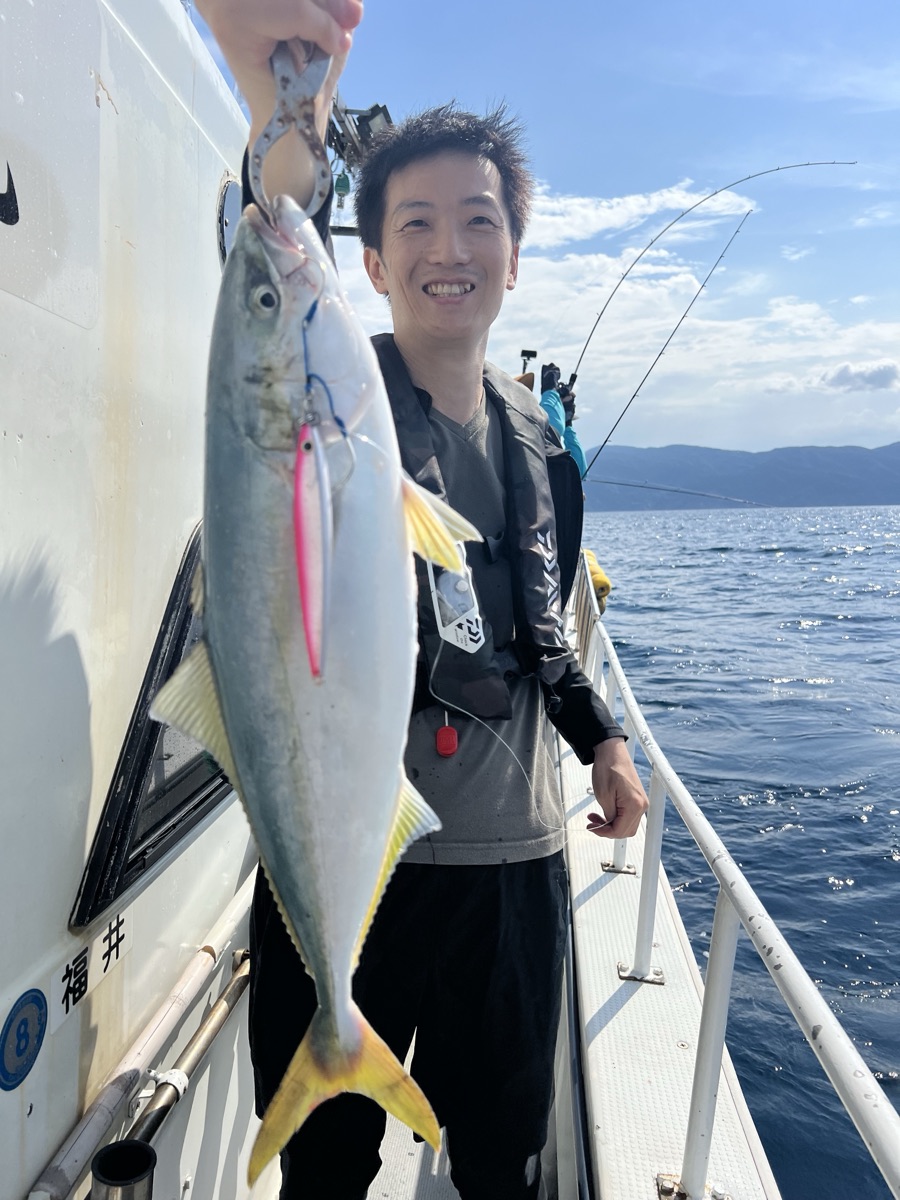 晴れた船上で、男性がジグで釣り上げた黄色い尾の魚を掲げて笑っている
