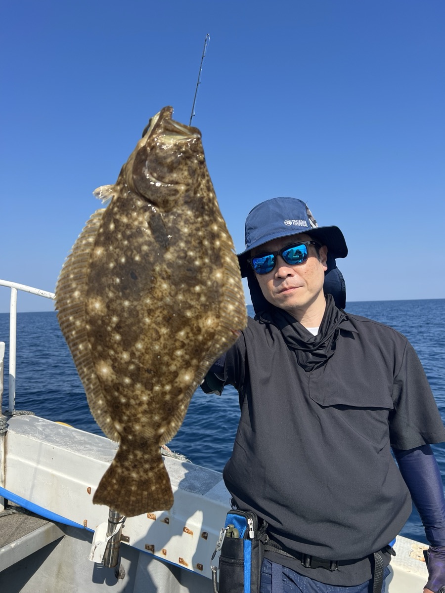 青空の船上で、サングラスをかけた男性が釣り上げた大きな平たい魚を掲げている
