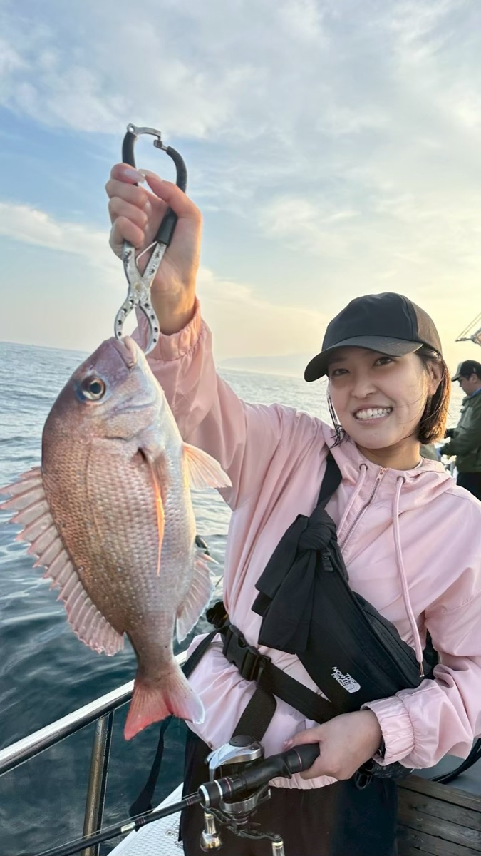 朝の船上で、ピンクの上着の女性が釣り上げた真鯛を掲げて笑っている